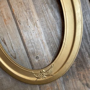 May include: Oval gold-colored picture frame with a decorative emblem at the bottom. The frame has a slightly textured surface and is set against a weathered wooden background. The emblem features a bow and banner design.