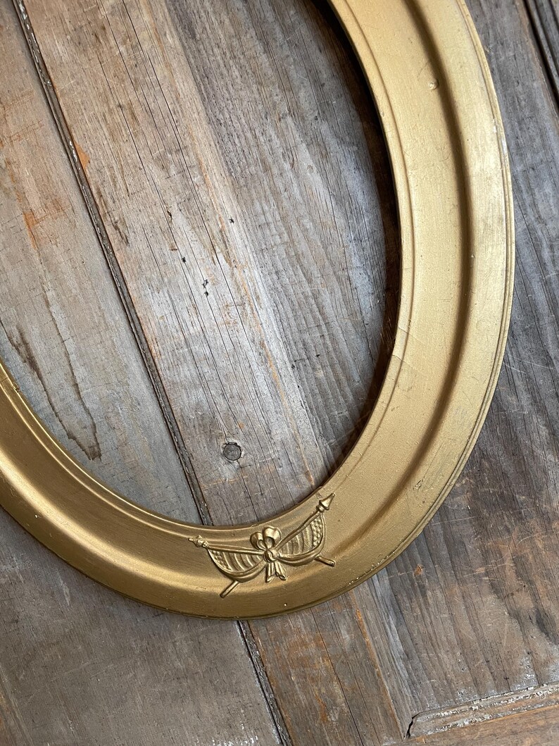 May include: Oval gold-colored picture frame with a decorative emblem at the bottom. The frame has a slightly textured surface and is set against a weathered wooden background. The emblem features a bow and banner design.