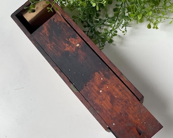 May include: A rectangular wooden box with a sliding lid featuring a dark, weathered brown finish with visible wood grain and small white speckles. The box has a hollow interior compartment at one end and a small notch on the lid for sliding.