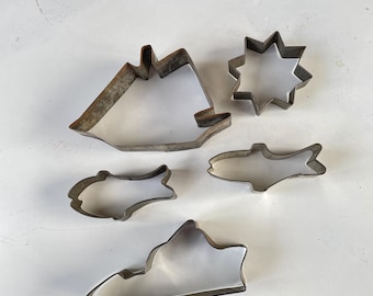 May include: A collection of vintage metal cookie cutters in various shapes, including a sailboat, star, fish, and mountain. The cutters are made of a dull, silver-colored metal and are arranged on a white surface. These are ideal for baking themed cookies.