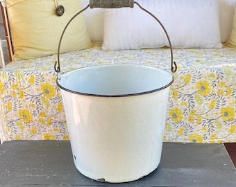 May include: A white enamel bucket with a dark rim and a metal handle featuring a wooden grip. The bucket has a cylindrical shape with a slightly tapered base and shows minor signs of wear along the bottom edge and the metal handle hardware.