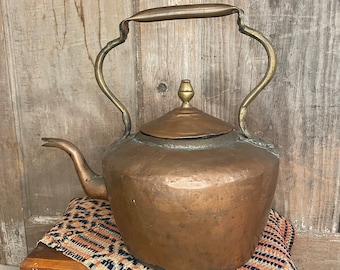 May include: A vintage copper kettle with a brass handle and spout. The kettle has a tarnished finish and is sitting on a patterned fabric.