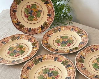 May include: A set of five vintage ceramic plates with a cream-colored base. Each plate features a central grape cluster design in shades of green, blue, and yellow, surrounded by a border of grape vines and leaves in brown and red.