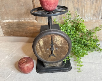 May include: An antique black metal scale with a round face and a decorative wheel-shaped platform. A red apple sits on the platform, and another rests on the surface below. The scale has the text "SARGENT & CO. New York, NY."