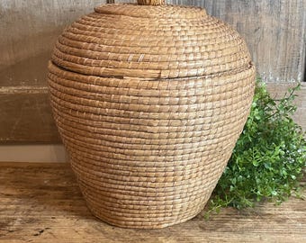 May include: A round, woven basket with a matching lid featuring a small, centered knob. The basket is constructed from light tan, natural fibers arranged in a tight, horizontal coil pattern, creating a sturdy, bulbous shape with a flat base.