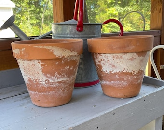 May include: Two terracotta flower pots with a weathered, white-washed finish on the exterior surface. Behind the pots sits a galvanized metal watering can featuring a bright red handle and a matching red rim at the base of the container.