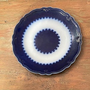 May include: A round, antique plate with a scalloped edge. The plate features a dark blue border and a white center with a blue, star-like pattern. The plate is set on a wooden surface.