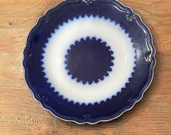 May include: A round, antique plate with a scalloped edge. The plate features a dark blue border and a white center with a blue, star-like pattern. The plate is set on a wooden surface.