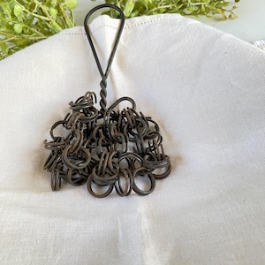 May include: A vintage metal kitchen whisk with a twisted handle and a cluster of small, interconnected rings. The whisk is dark brown and rests on a white cloth, with green foliage in the background. This rustic kitchen tool is designed for whisking ingredients.
