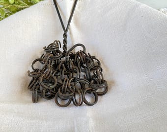 May include: A vintage metal kitchen whisk with a twisted handle and a cluster of small, interconnected rings. The whisk is dark brown and rests on a white cloth, with green foliage in the background. This rustic kitchen tool is designed for whisking ingredients.