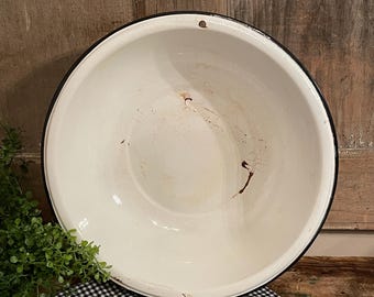 May include: A round white enamel basin with a black rim. The interior surface features visible signs of wear, including brown staining and small chips in the enamel coating. A single small hole is positioned near the top edge of the basin rim.