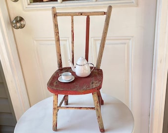May include: A small, weathered wooden chair with a distressed red painted seat and natural wood finish on the legs and backrest. Resting on the seat is a miniature white ceramic teapot and a matching teacup on a small saucer with silver trim.