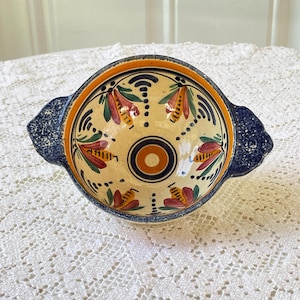 May include: A ceramic bowl with two handles, featuring a cream-colored interior decorated with hand-painted red and green floral motifs and a central orange circle. The exterior handles are dark blue with a speckled white texture.