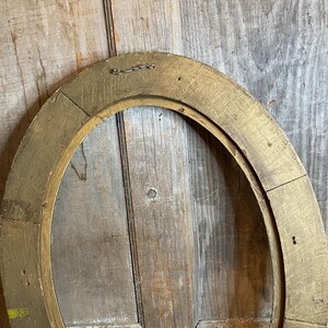 May include: Oval-shaped, antique gold picture frame. The frame is made of wood and has a distressed, aged appearance. The frame is empty, revealing the wooden backing. A small metal wire is attached to the top for hanging.