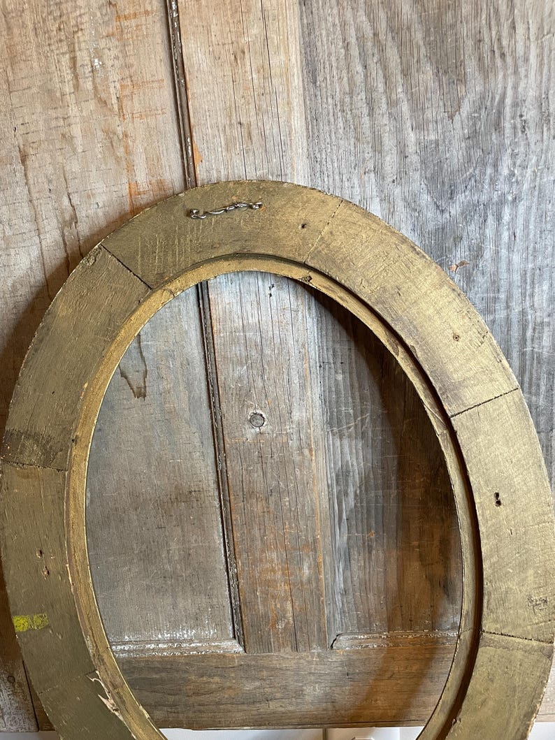 May include: Oval-shaped, antique gold picture frame. The frame is made of wood and has a distressed, aged appearance. The frame is empty, revealing the wooden backing. A small metal wire is attached to the top for hanging.