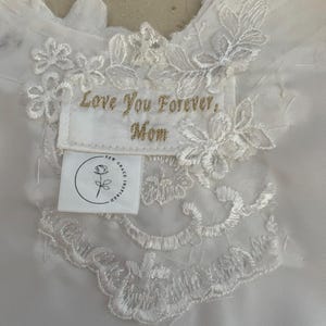 May include: White fabric with intricate lace detailing and a sewn-on label. The label features gold embroidered text reading 'Love You Forever, Mom.' Below the text is a small white tag with a rose logo and the words 'Sew Grace Inspired.' The lace design includes floral and leaf patterns.