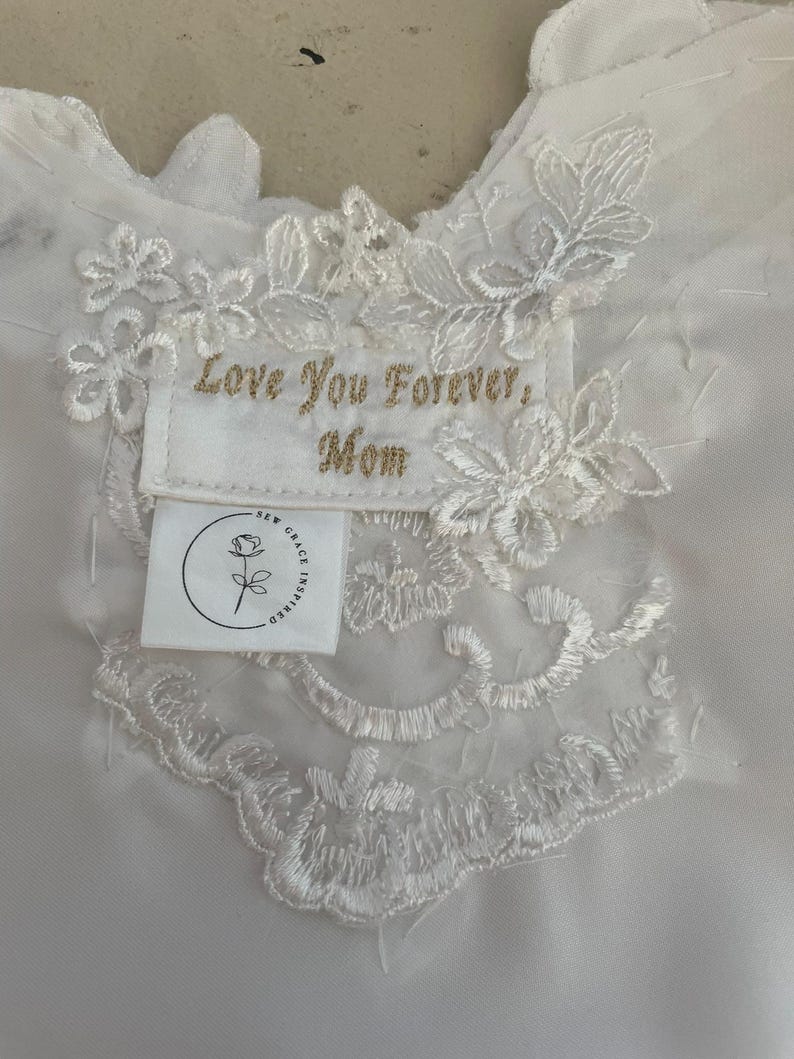 May include: White fabric with intricate lace detailing and a sewn-on label. The label features gold embroidered text reading 'Love You Forever, Mom.' Below the text is a small white tag with a rose logo and the words 'Sew Grace Inspired.' The lace design includes floral and leaf patterns.