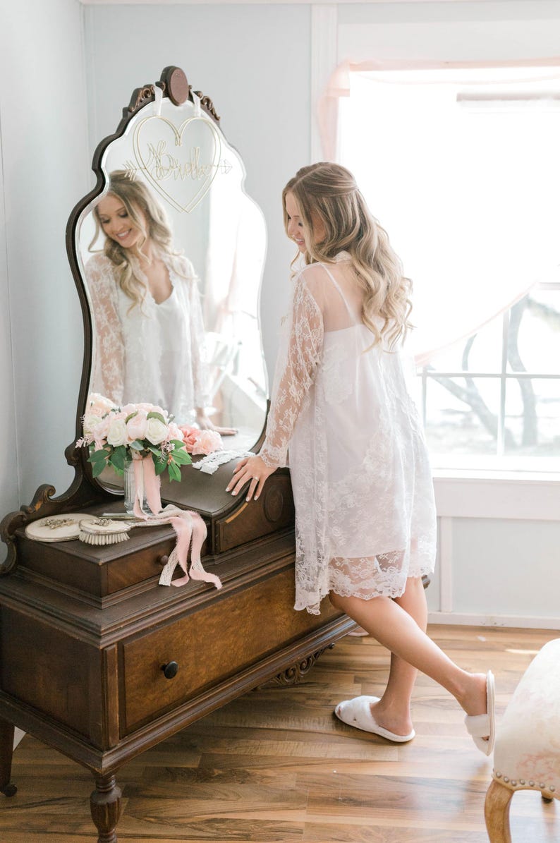 May include: A woman in a white lace robe stands near a wooden vanity with a large mirror. The mirror has a heart-shaped design with the word 'Bride' on it. A bouquet of flowers and a brush sit on the vanity. The room has light blue walls and a wooden floor.