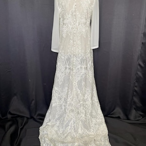 May include: A white wedding dress with intricate lace detailing. The dress features long, sheer sleeves and a high neckline. The lace design extends from the bodice to the skirt, which flares out at the bottom.