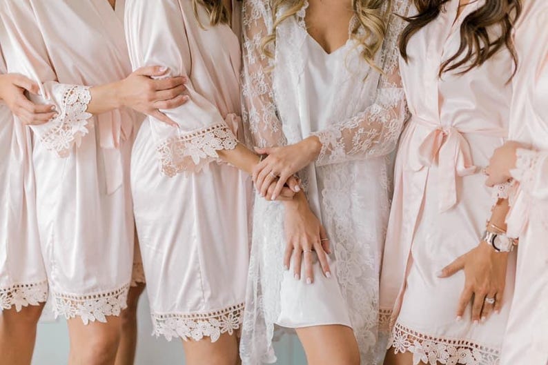 May include: A group of women wearing matching light pink satin robes with lace trim. One woman wears a white lace robe over a white slip. The robes have a tie closure and are knee-length. The lace trim is around the sleeves and hem.