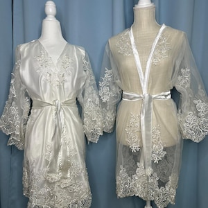 May include: Two white bridal robes displayed on mannequins. The robe on the left is a satin fabric with lace detailing on the sleeves and hem, and a matching tie. The robe on the right is sheer with lace accents and a satin tie. Both robes are elegant and suitable for a wedding.
