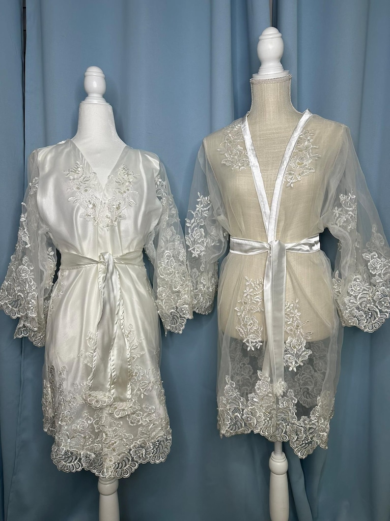 May include: Two white bridal robes displayed on mannequins. The robe on the left is a satin fabric with lace detailing on the sleeves and hem, and a matching tie. The robe on the right is sheer with lace accents and a satin tie. Both robes are elegant and suitable for a wedding.