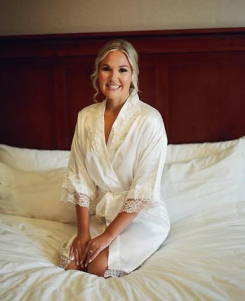 May include: A woman wearing a white satin robe with lace trim, kneeling on a white bed. The robe has a wrap-around design with a tie at the waist. The woman has blonde hair and is smiling. The background is a dark wooden headboard.