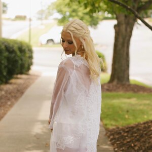 May include: A white, sheer, lace-detailed dress. The dress has a long, flowing design with intricate lace patterns. The dress is worn over a white slip, and the overall look is elegant and ethereal. The model is standing on a sidewalk with a green lawn and trees in the background.