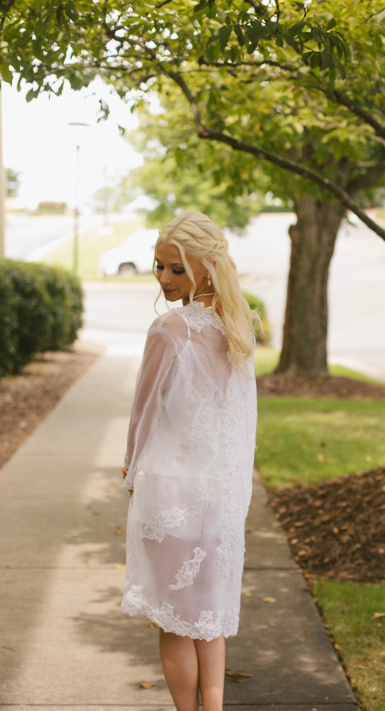 May include: A white, sheer, lace-detailed dress. The dress has a long, flowing design with intricate lace patterns. The dress is worn over a white slip, and the overall look is elegant and ethereal. The model is standing on a sidewalk with a green lawn and trees in the background.