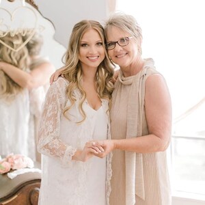 May include: Two women smiling, embracing, and holding hands. The younger woman wears a white lace jacket over a white dress. The older woman wears a beige dress and glasses. A mirror with a heart-shaped decoration is visible in the background.