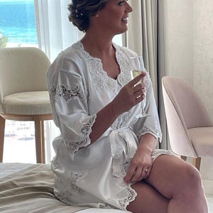 May include: A white satin robe with delicate lace trim. The robe has a wrap-around design with a matching tie. The lace detailing is present on the sleeves and along the edges of the robe. The woman is holding a champagne flute, suggesting a celebratory occasion.