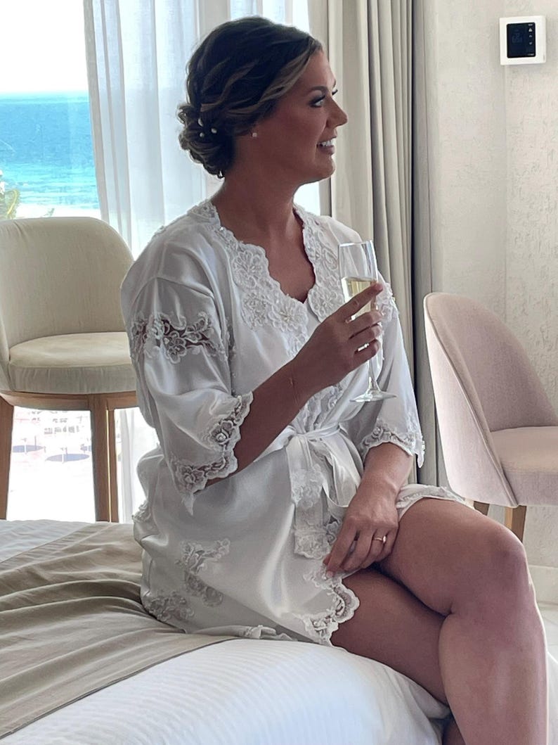 May include: A white satin robe with delicate lace trim. The robe has a wrap-around design with a matching tie. The lace detailing is present on the sleeves and along the edges of the robe. The woman is holding a champagne flute, suggesting a celebratory occasion.