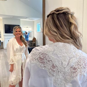 May include: A bride-to-be in a white satin robe with lace trim, standing in front of a mirror. The robe is tied at the waist and has a decorative lace design on the back. The bride's hair is styled in a half-up, half-down style with a braided crown. The mirror reflects the room, including a person in a blue shirt.