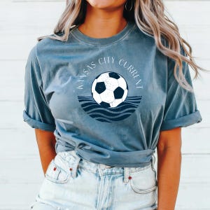 May include: A blue-grey t-shirt featuring a soccer ball design with the text "Kansas City Current" in a circular arrangement. The soccer ball is black and white, set above blue wave graphics.