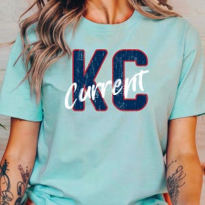 May include: A light blue t-shirt with the text "KC Current" in a distressed navy blue and red design. The shirt is a solid color and has short sleeves.