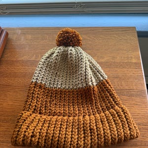 May include: A two-toned knit beanie featuring a tan upper section and a dark orange lower section with a ribbed cuff. The hat is topped with a matching dark orange pom-pom, showcasing a textured, chunky yarn construction throughout the entire piece.