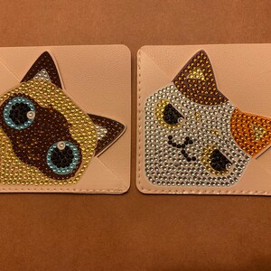 May include: Two square coasters with cat faces. One coaster features a Siamese cat face with brown, gold, and blue rhinestones. The other coaster has a calico cat face with brown, silver, and orange rhinestones. Both coasters have a light tan background.