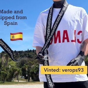 Könnte beinhalten: Ein weißes kurzärmeliges T-Shirt mit großen roten Buchstaben auf der Brust und einem langen schwarzen Stoffband, das um den Hals drapiert ist. Das schwarze Band ist mit wiederholtem weißem Text bedruckt und hängt vorne am T-Shirt herab.