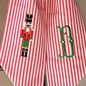 Puede incluir: Decoración navideña de tela a rayas rojas y blancas con un cascanueces y un monograma verde. La tela cuelga de una guirnalda verde. El cascanueces está bordado en rojo, verde, negro y dorado.