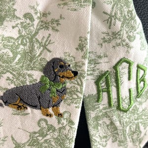 May include: A cream-colored fabric ribbon featuring a green toile de jouy pattern, an embroidered gray and tan dachshund wearing a green bow, and the embroidered green monogram letters ACB in a decorative font on the right side of the ribbon.