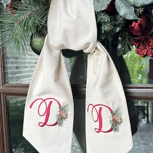 Puede incluir: Bufanda color crema con una letra "D" bordada en rojo y detalles de piñas y vegetación. La bufanda está colocada frente a una corona navideña con adornos rojos y verdes.