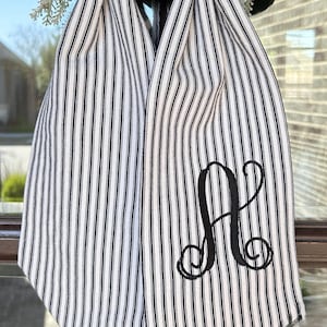 May include: A black and white striped scarf with a large embroidered black letter "A". The scarf is made of a soft fabric and has a classic, timeless design. The scarf is hanging in front of a window.