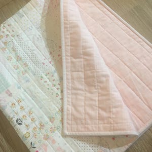 May include: A handmade baby quilt with a patchwork design. The quilt features pastel fabrics, including floral prints, polka dots, and geometric patterns. The reverse side is a solid light pink fabric. The quilt is machine quilted with a stitched border.
