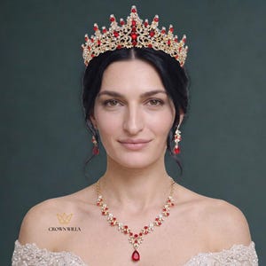 Può includere: Una tiara color oro con un intricato lavoro in metallo, cristalli trasparenti e pietre rosse a forma di goccia. Il set include orecchini pendenti abbinati e una collana con un ciondolo centrale a goccia rossa. Il testo CROWN WILLA è visibile.