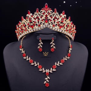Op de afbeelding: Een goudkleurige sieradenset bestaande uit een tiara, een ketting en bijpassende oorbellen. De tiara is versierd met talloze rode druppelvormige en ronde kristallen. De ketting en oorbellen hebben een delicaat rankmotief met rode bloemen.