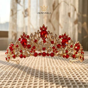 May include: A gold-toned metal tiara featuring a symmetrical filigree design adorned with numerous small clear crystals and several prominent, teardrop-shaped and round red gemstones. The text CROWN WILLA is printed above the tiara.