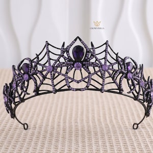 May include: A black metal tiara featuring a spider web design adorned with purple rhinestones and larger purple teardrop and round crystals. The piece includes spider motifs and the text CROWN WILLA printed in gold near the top centre.