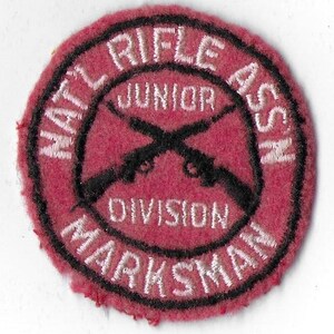 Puede incluir: Un parche de fieltro rojo con bordados en blanco y negro. El parche dice "National Rifle Association Junior Division Marksman".