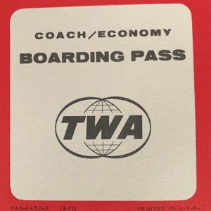 May include: A vintage TWA boarding pass with the text "COACH/ECONOMY BOARDING PASS" in black. The TWA logo is centered, with the text "TWA" inside a double oval with a globe design. The pass is on a red background.