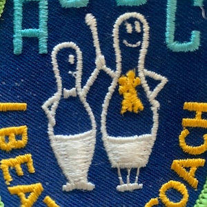 May include: Embroidered patch with a blue background and a green border. The patch features two white cartoon figures with arms raised, and the text "I BEAT MY COACH" and "AJBC" in yellow and light blue.